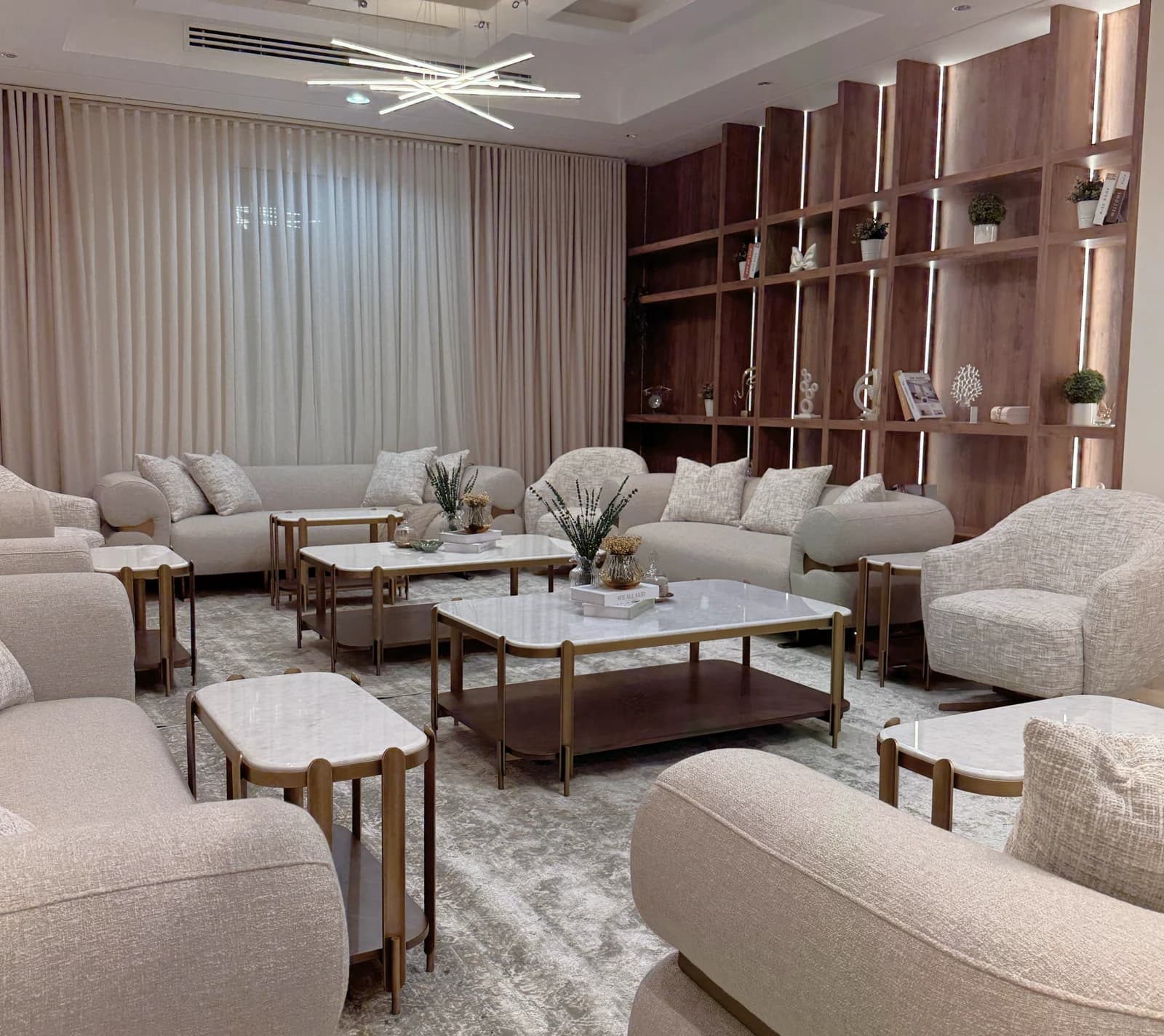 A modern living room features plush beige sofas and armchairs, a geometric chandelier, marble-top coffee tables, and an elegant wooden shelving unit adorned with small plants and decor.